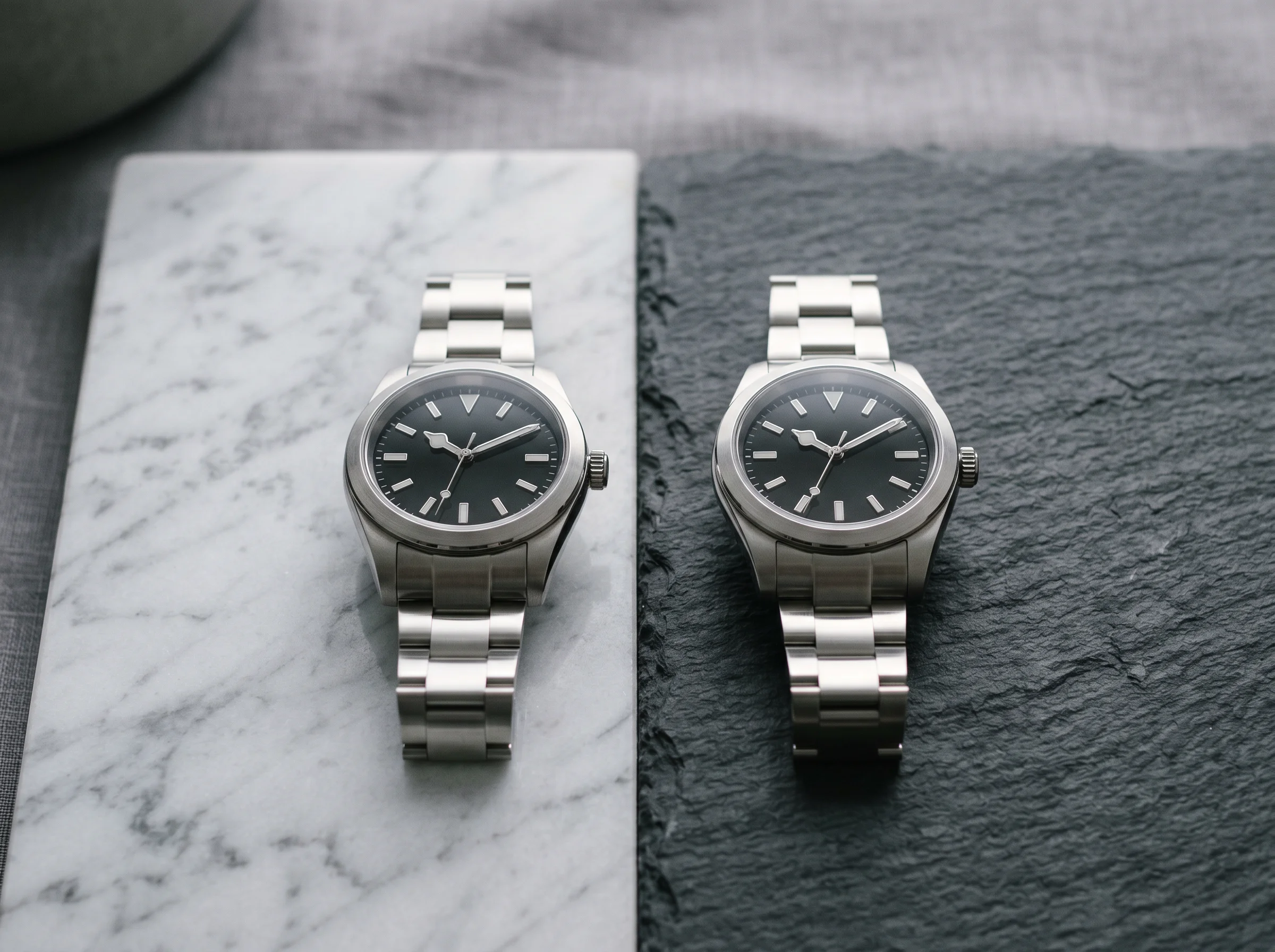 Two similar unbranded steel sport watches side by side on a split marble and slate surface, suggesting a comparison