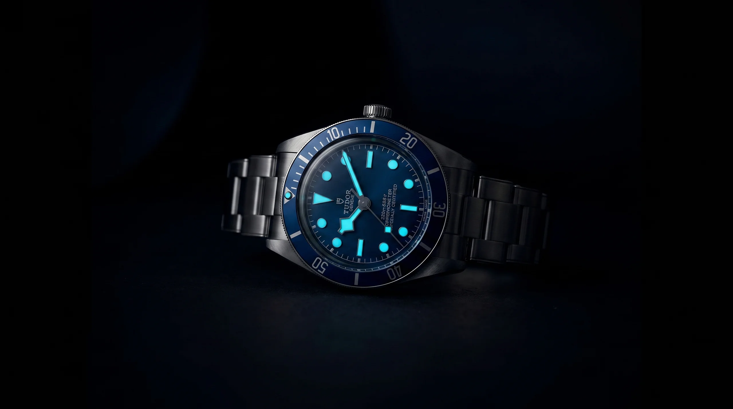 A Tudor Black Bay dive watch glowing blue-green in the dark, showing Super-LumiNova at work