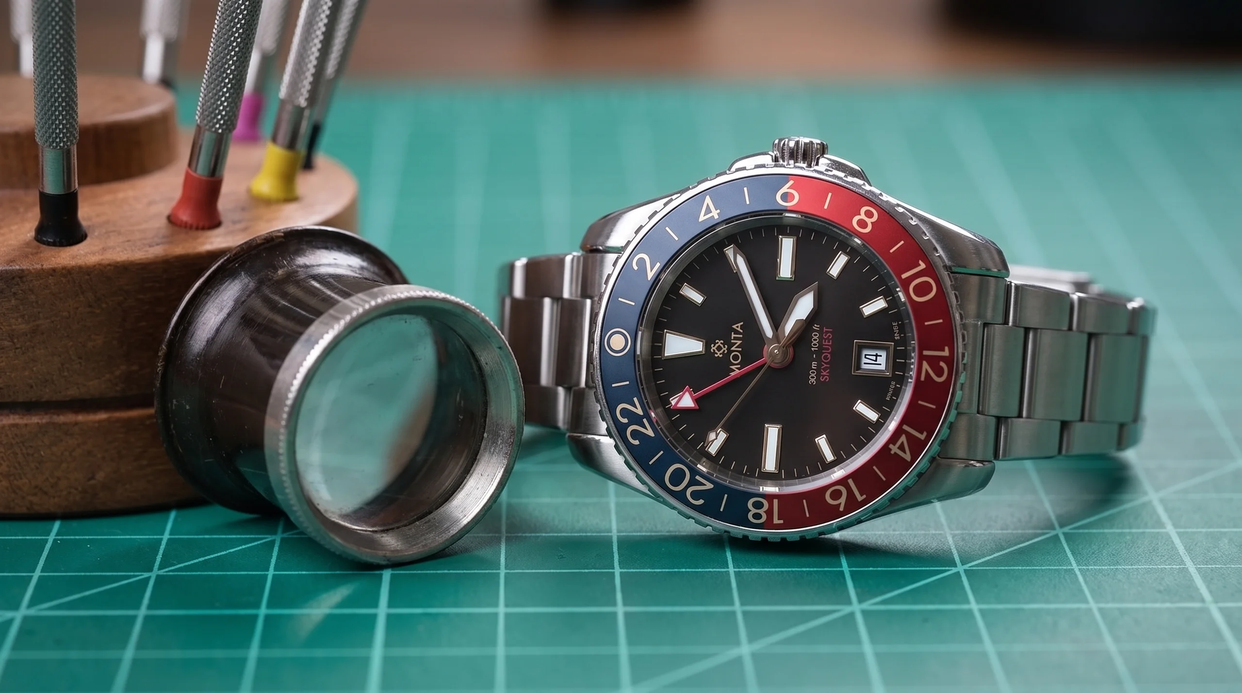 Watchmaker's loupe and screwdriver set beside a Monta Skyquest GMT watch on a green cutting mat workbench