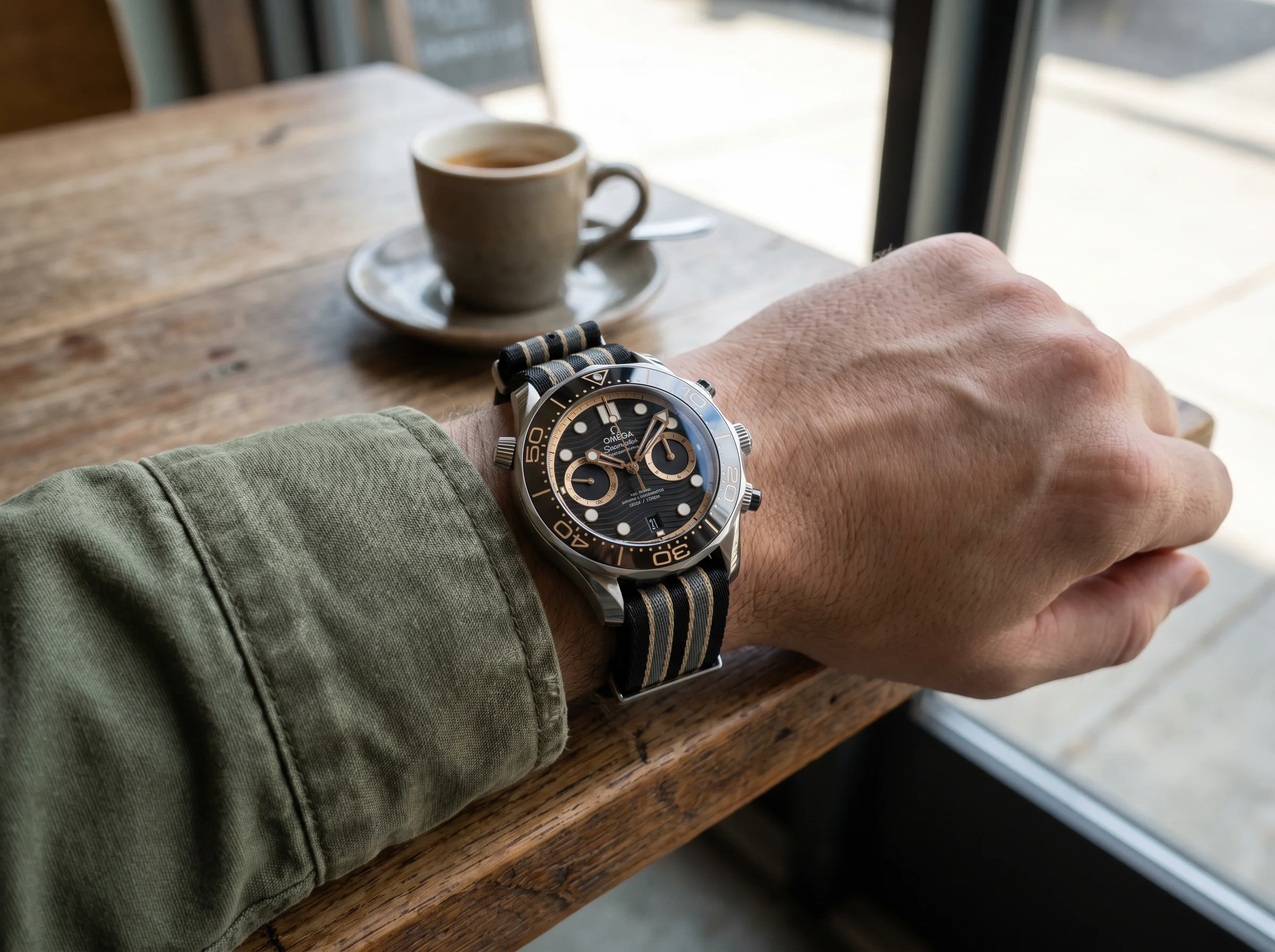 Omega Seamaster 007 First Light worn on a wrist over an olive field jacket cuff at a cafe table