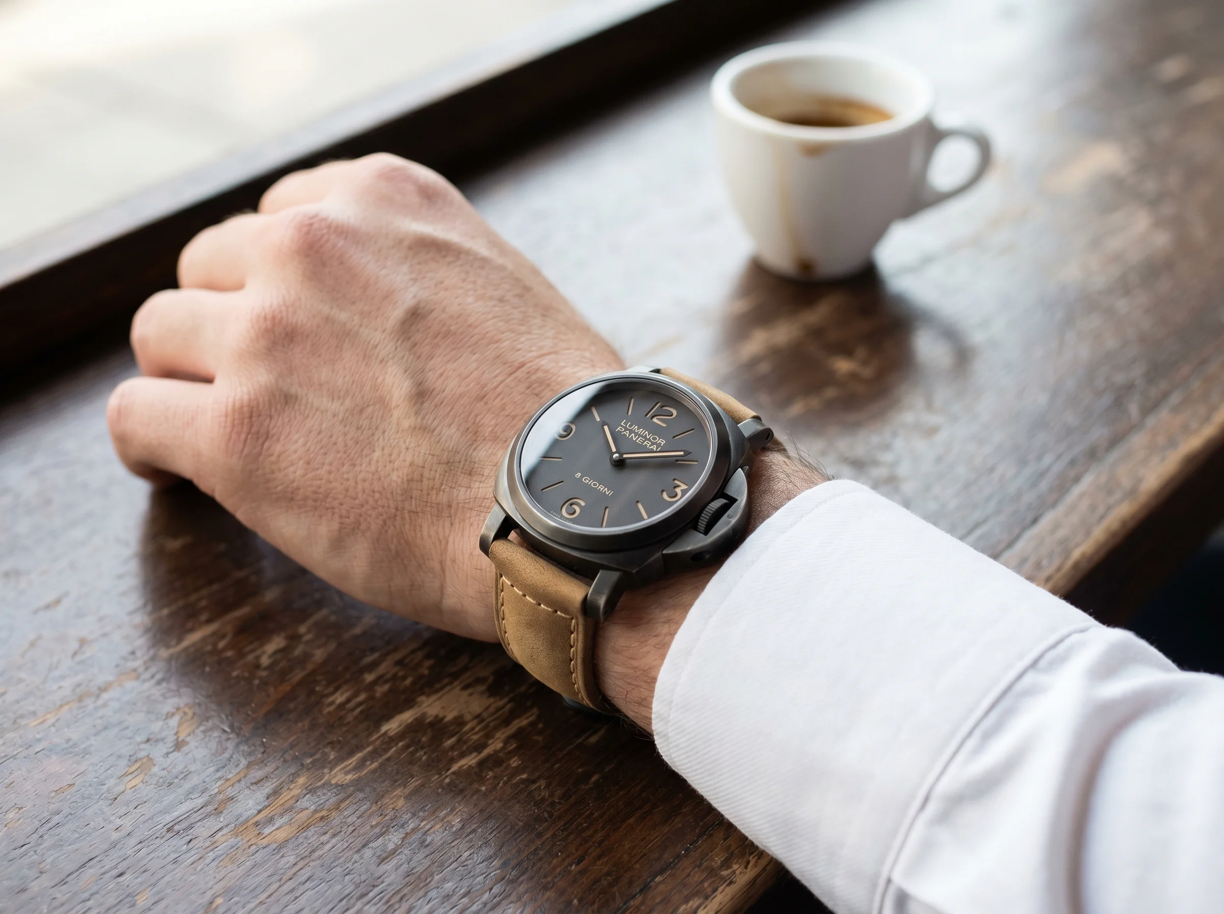 Panerai Luminor 8 Giorni Brunito PAM01733 worn on a man's left wrist resting on a dark wood cafe table with a small white porcelain espresso cup in soft focus and a white dress shirt cuff visible at the frame edge in gentle morning daylight