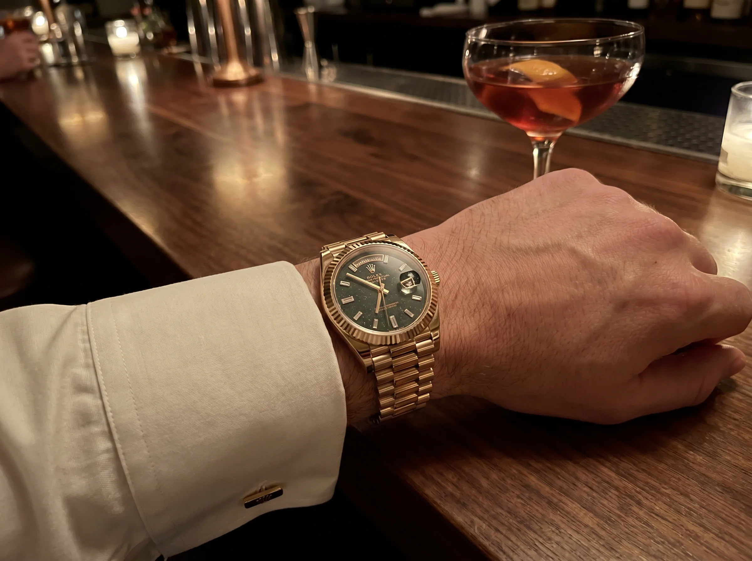 Wrist shot of a Rolex Day-Date 40 reference 228235JG-0003 in Jubilee Gold worn on the left wrist at a dim evening wine bar, with a Negroni in the background and white dress shirt cuff at the lower frame edge.
