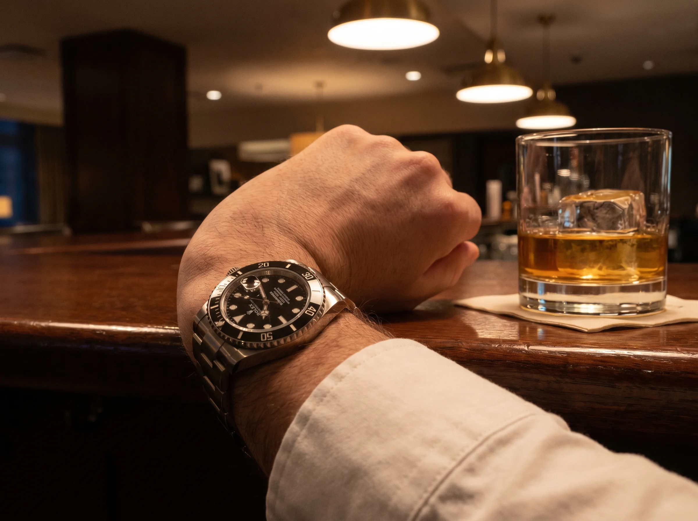 Wrist shot of a Rolex Submariner Date reference 126610LN black-on-black current production model at a hotel lounge bar with a tumbler of whiskey, the standard steel Submariner that absorbs displaced demand from the Cookie Monster discontinuation.
