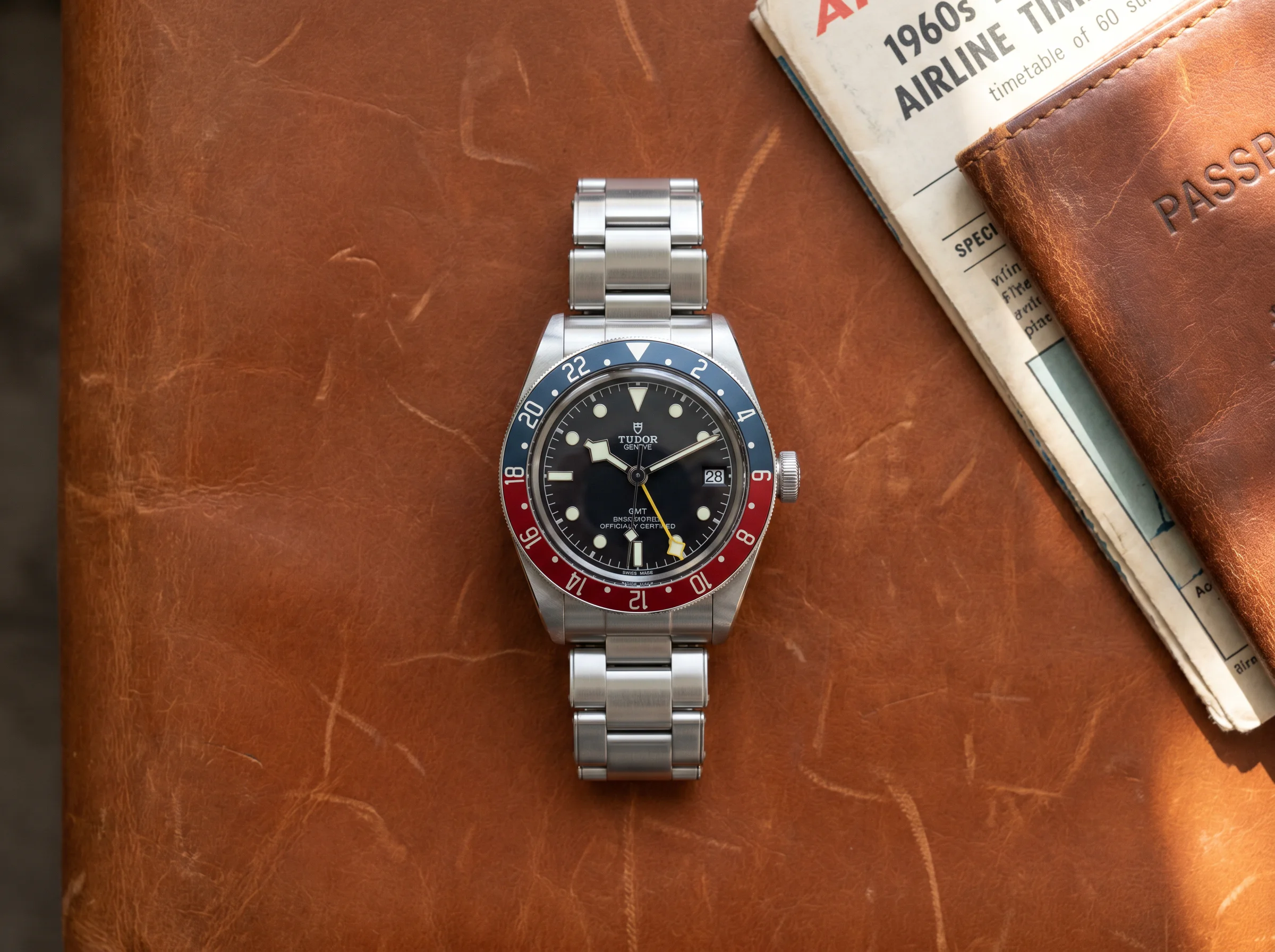 Tudor Black Bay GMT 79830RB with blue and red Pepsi aluminum bezel and yellow snowflake-tipped GMT hand, top-down on cognac leather travel portfolio